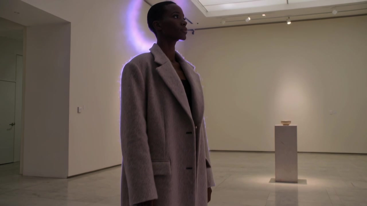 Uscendo da una porta a sinistra, una donna in cappotto lungo cammina sul pavimento della galleria, osservando una ciotola illuminata su un piedistallo.