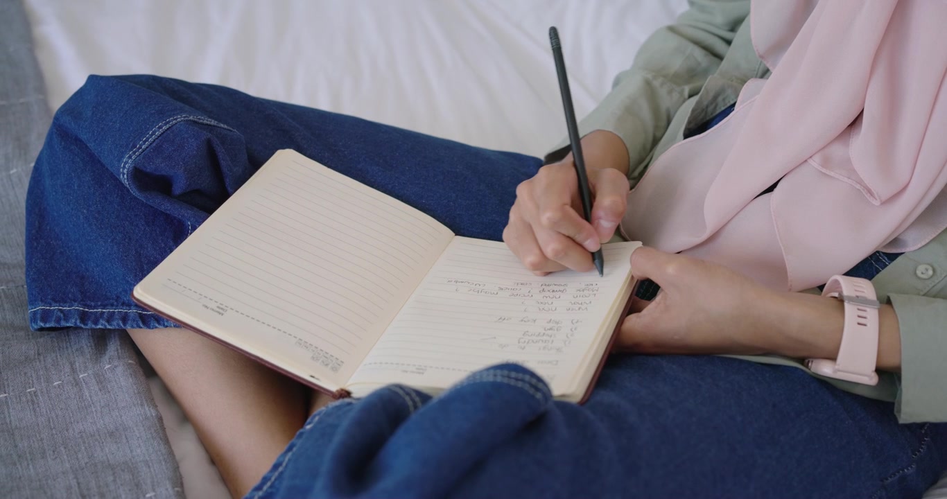 Mano derecha bajando un lápiz sobre una página, escribiendo una lista en un cuaderno abierto sobre la cama para planificar