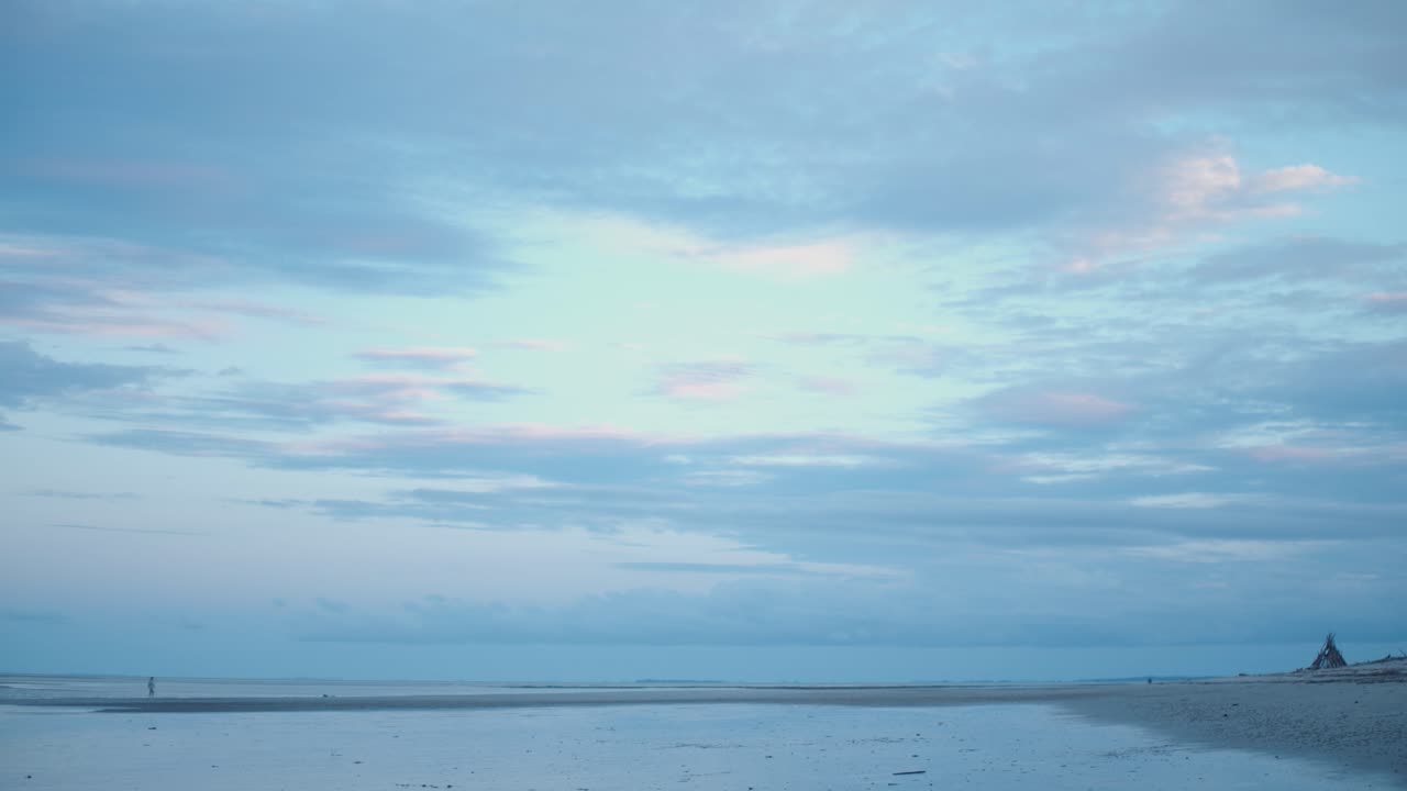 한 소녀가 자전거를 타고 막대기로 만든 천막을 지나 반사되는 해변을 가로질러 지는 태양이 오후의 푸른 시간에 후퇴하는 물과 푹신한 분홍색 구름에 시원한 색을 쏟습니다.