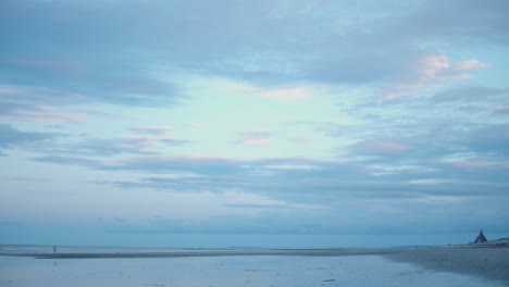 una niña monta su bicicleta pasando un tipi hecho de palos y cruzando la playa reflectante mientras el sol poniente derrama colores fríos en el agua que se retira y nubes rosas esponjosas a la hora azul de la tarde
