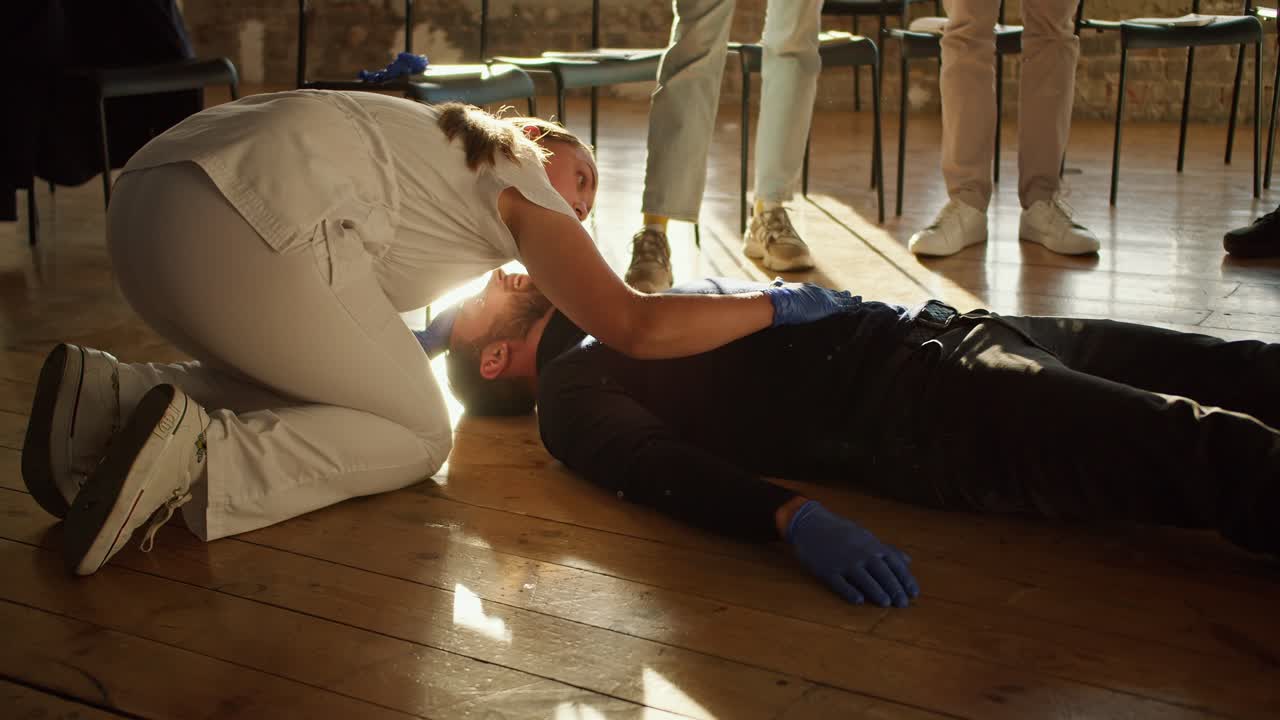 클로즈업 촬영: 색 의료 유니폼을 입은 전문 간호사 소녀가 한 학생의 호흡을 확인하기 위해 한 무리의 사람들을 훈련시니다.