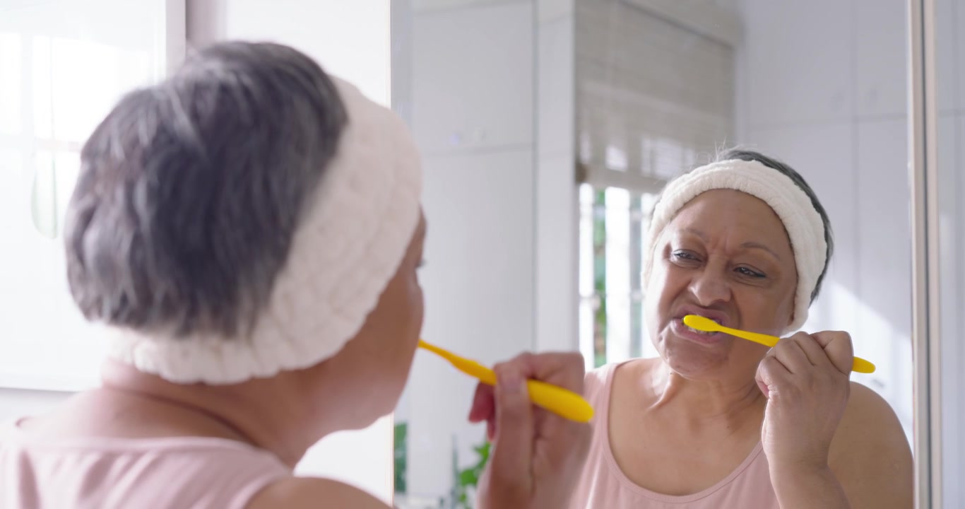 Senior African woman checking mirror grabbing yellow brush brushing teeth at vanity for oral care