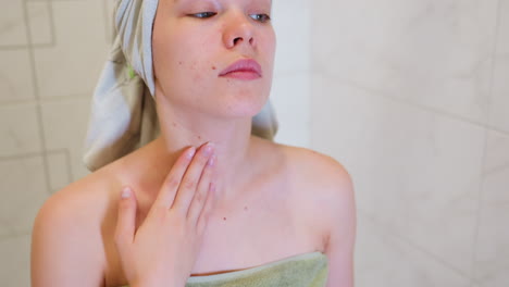 Close-up view of woman wrapped in towel, with hair wrapped in towel, applying cream on her neck, she is focused on moisturizing and caring for her skin after shower, enhancing relaxation