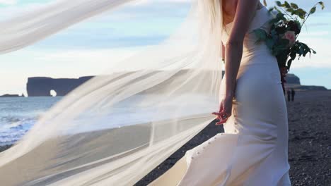 long wedding bridal veil flowing in the wind behind caucasian bride in white dress standing on black sand beach during sunset