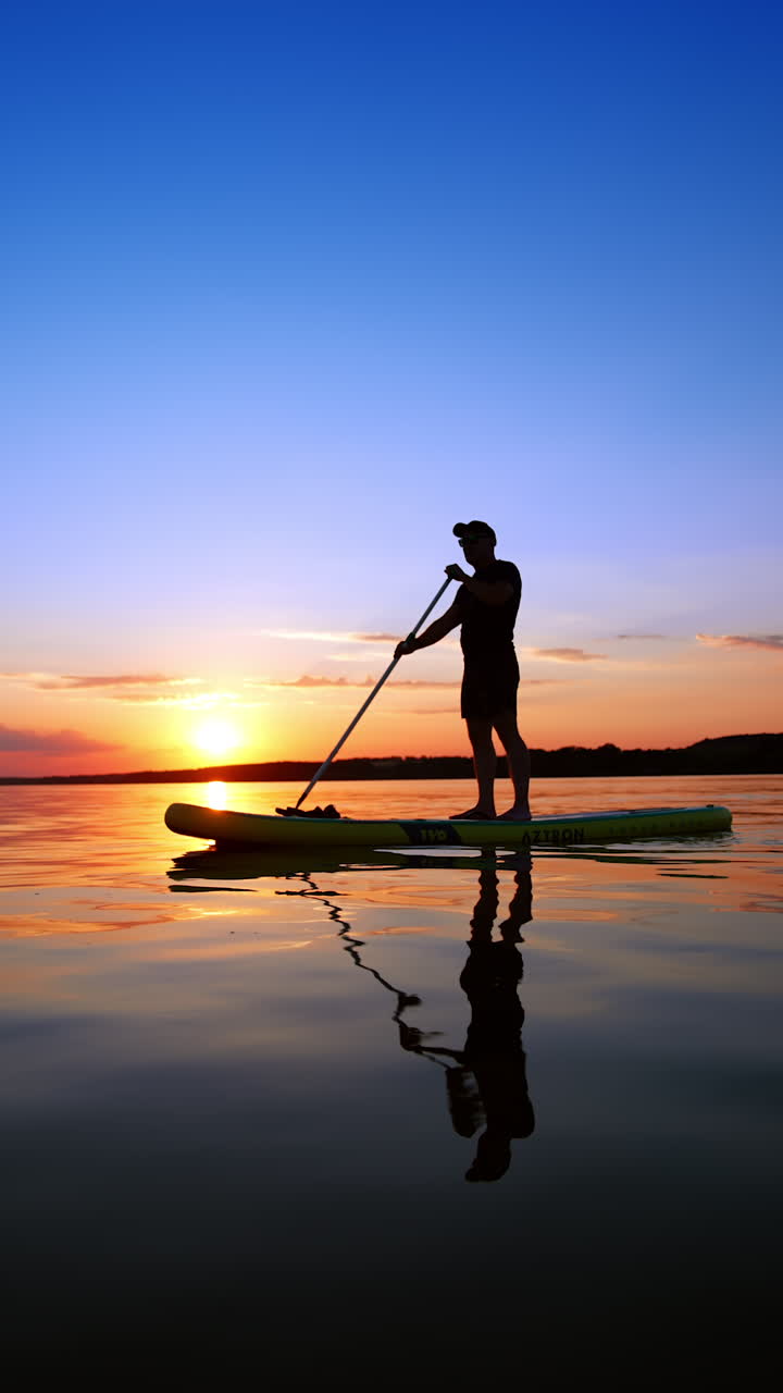 Sup boarding training on the river at sunset. Man stands confidently rowing with an oar. Low angle view. Vertical video.