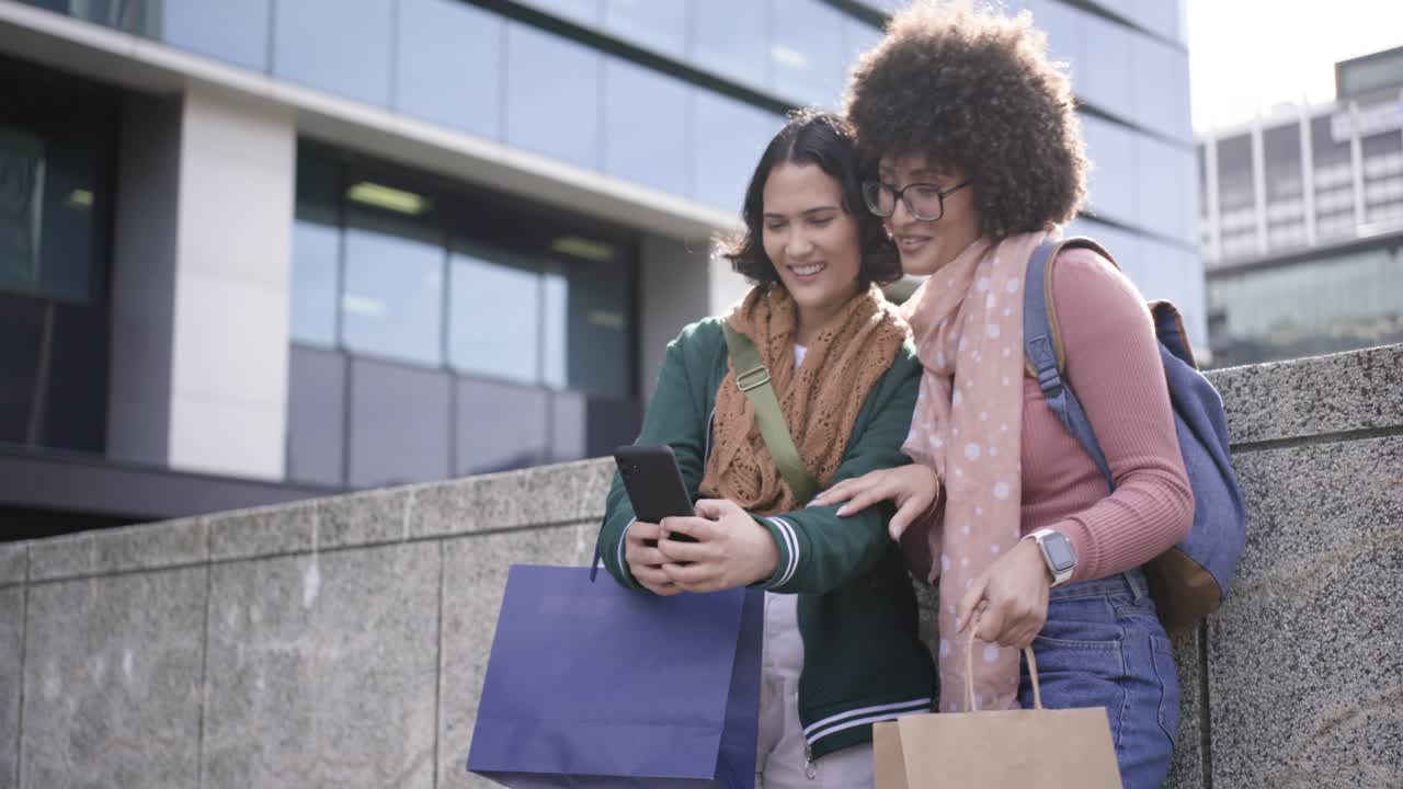 Amiche con borse che mostrano il contenuto dello smartphone, appoggiate al bordo di una piazza e che festeggiano i loro acquisti.