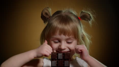 niña pequeña niño comiendo postre de barra de chocolate, mostrando los pulgares hacia arriba aislados en fondo oscuro