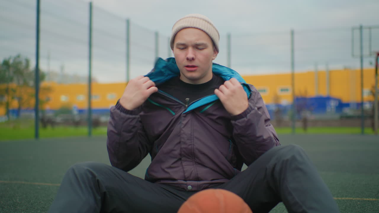 Giovane uomo bianco seduto su un campo da basket con indosso un berretto e una giacca, che tiene in mano un pallone mentre si riposa dopo l'allenamento, parco cittadino recintato sullo sfondo con magazzino giallo e cielo nuvoloso, scarpe da ginnastica e manto erboso