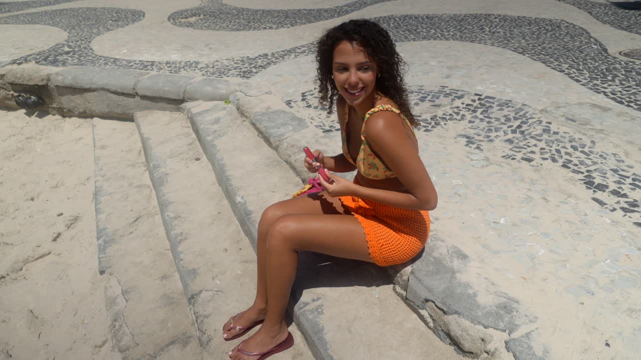 Giovane donna brasiliana di colore si sta mettendo il rossetto mentre è seduta sui gradini sul marciapiede di Copacabana a Rio de Janeiro, Brasile, in una giornata di sole. Statico, medio in 4K.
