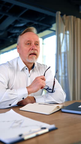 pensive fair-haired mature man sits at desk holding glasses at his mouth. man distracts from his thoughts abruptly. low angle view. vertical video.