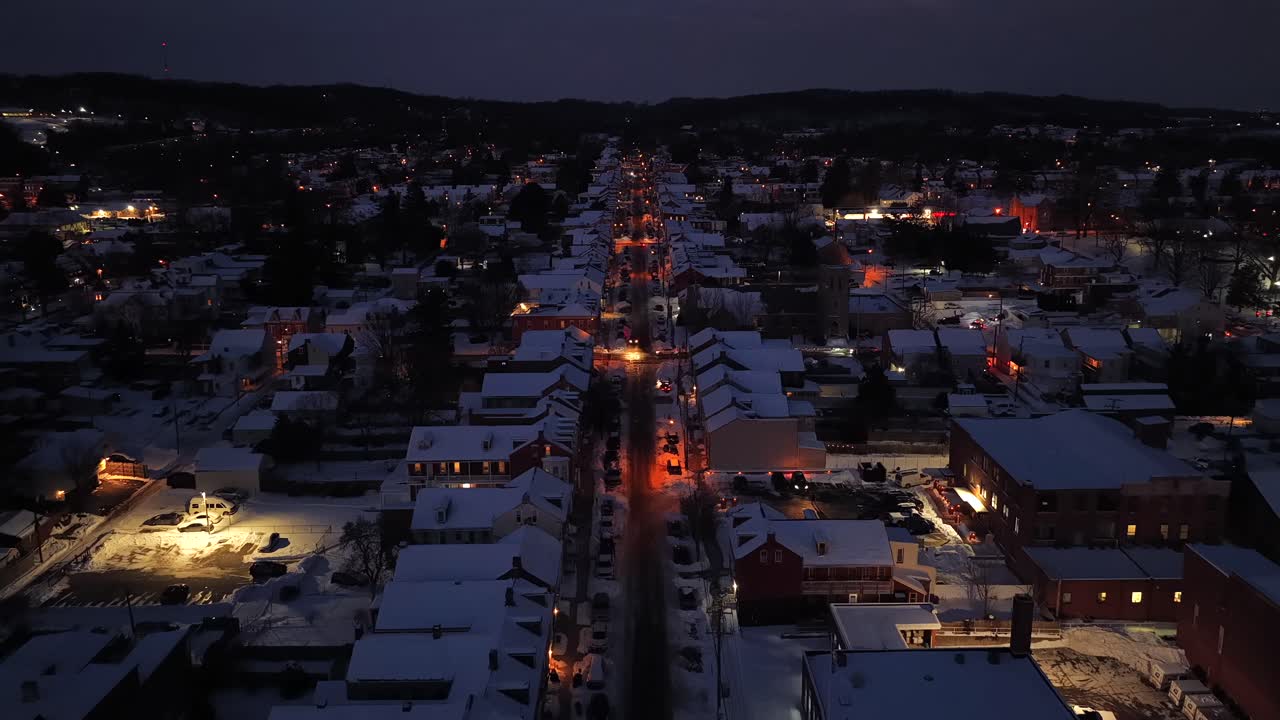 Hidas drone-lento valaistun, viihtyisän pienen amerikkalaisen kaupungin pääkadun yllä talvella. Lumiset rivitalojen katot tien varrella. Ilmakuva ylilennosta. Joulun aikaan yöllä Yhdysvalloissa. Rauhallinen kaupunki