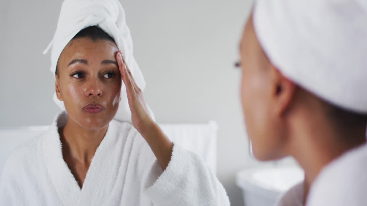 African american woman in bathrobe applying face cream looking in the mirror at bathroom