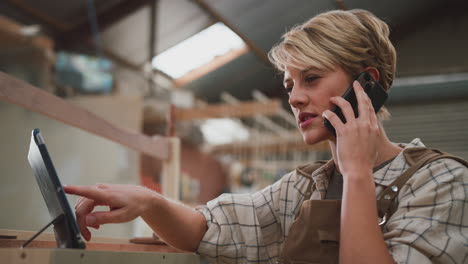 female apprentice carpenter with digital tablet working in furniture workshop making phone call