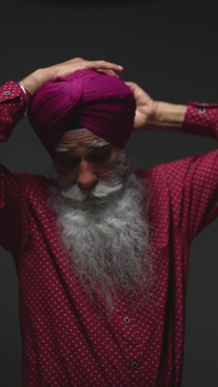 video vertical de iluminación de estudio de baja clave de un hombre sikh anciano con barba atando tela para turbante contra un fondo oscuro 5