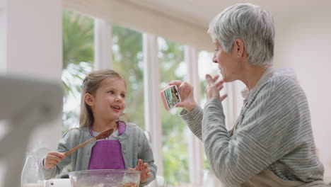 Glückliche-Großmutter-Fotografiert-Mit-Dem-Smartphone-Die-Süße-Enkelin-In-Der-Küche-Und-Mischt-Zutaten-Zum-Backen,-Schmeckt-Schokoladensoße.-Oma-Teilt-Ihr-Wochenende-Mit-Ihrem-Enkelkind-In-Den-Sozialen-Medien