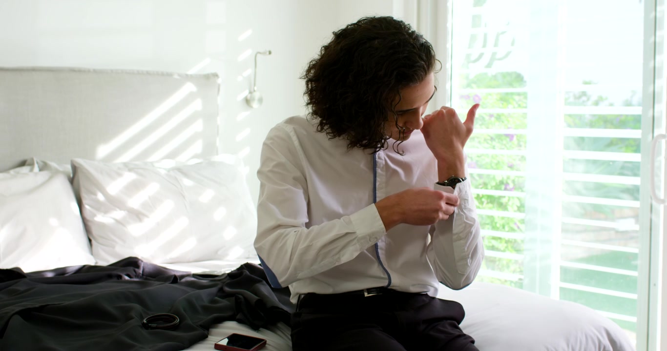After checking watch African American man sitting on bed adjusting strap and shirt cuff before work
