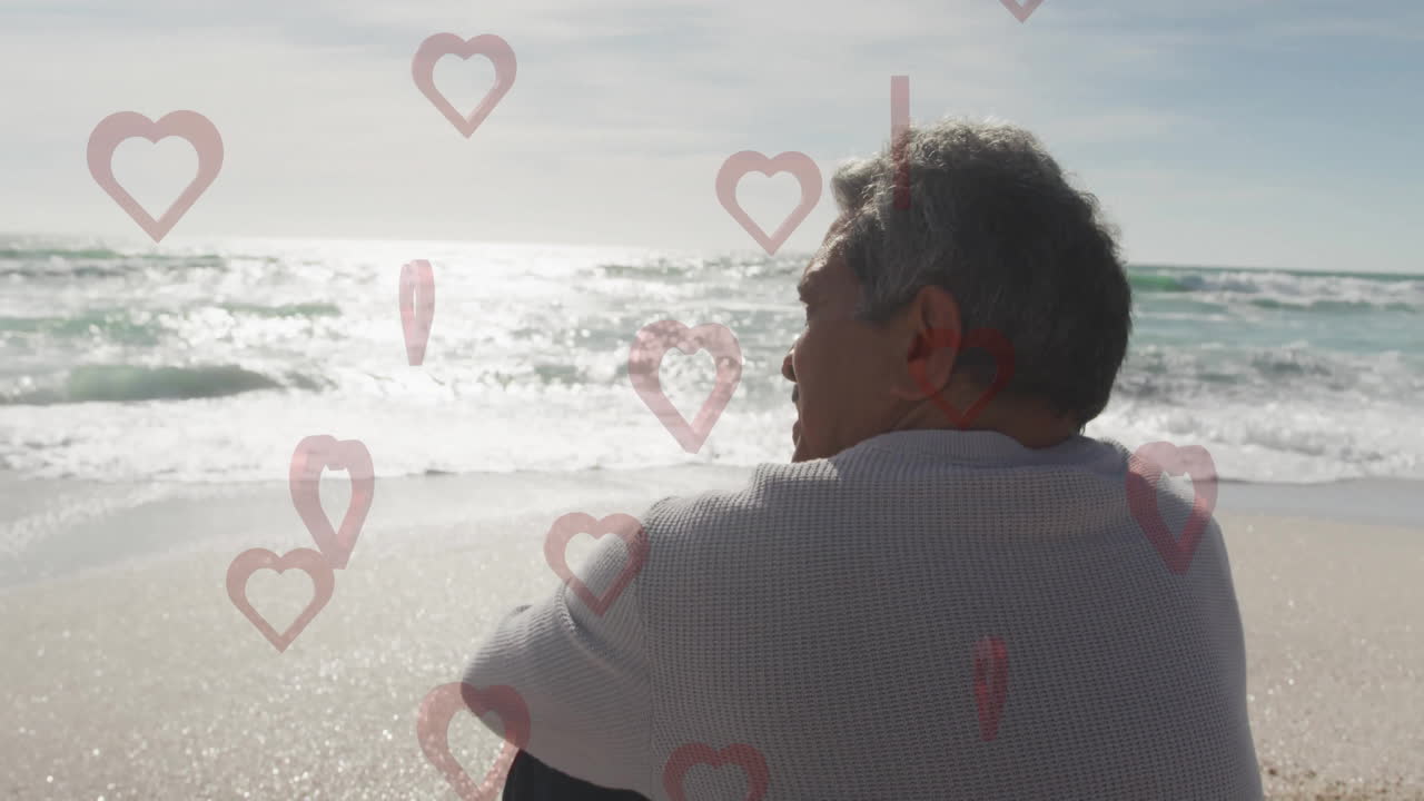 On beach, older man sitting with animation of heart icons surrounding him
