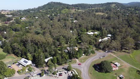 Aerial-view-of-a-green-Australian-countryside-flying-over-road,-roundabout-towards-a-green-hill-dotted-with-private-homes