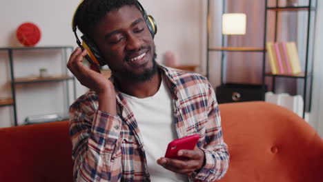 African-American-young-man-in-headphones-listening-music-dancing,-singing-in-living-room-at-home