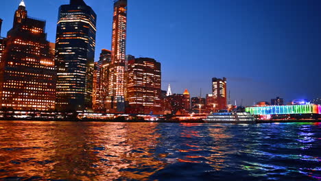 Luminous skyline of modern New York reflects in the waterscape of the East River. Multi-deck cruise boat stands near the Pier 17 Bar