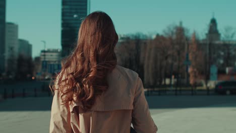 camera follows beautiful woman with long curly brown hair in autumn clothes walk alone on empty city square slow motion.