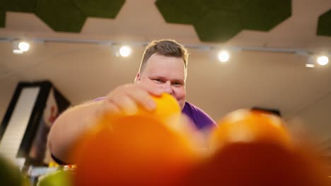 Shot-from-the-counter-of-a-happy-overweight-man-in-a-purple-T-shirt-choosing-oranges-from-the-counter