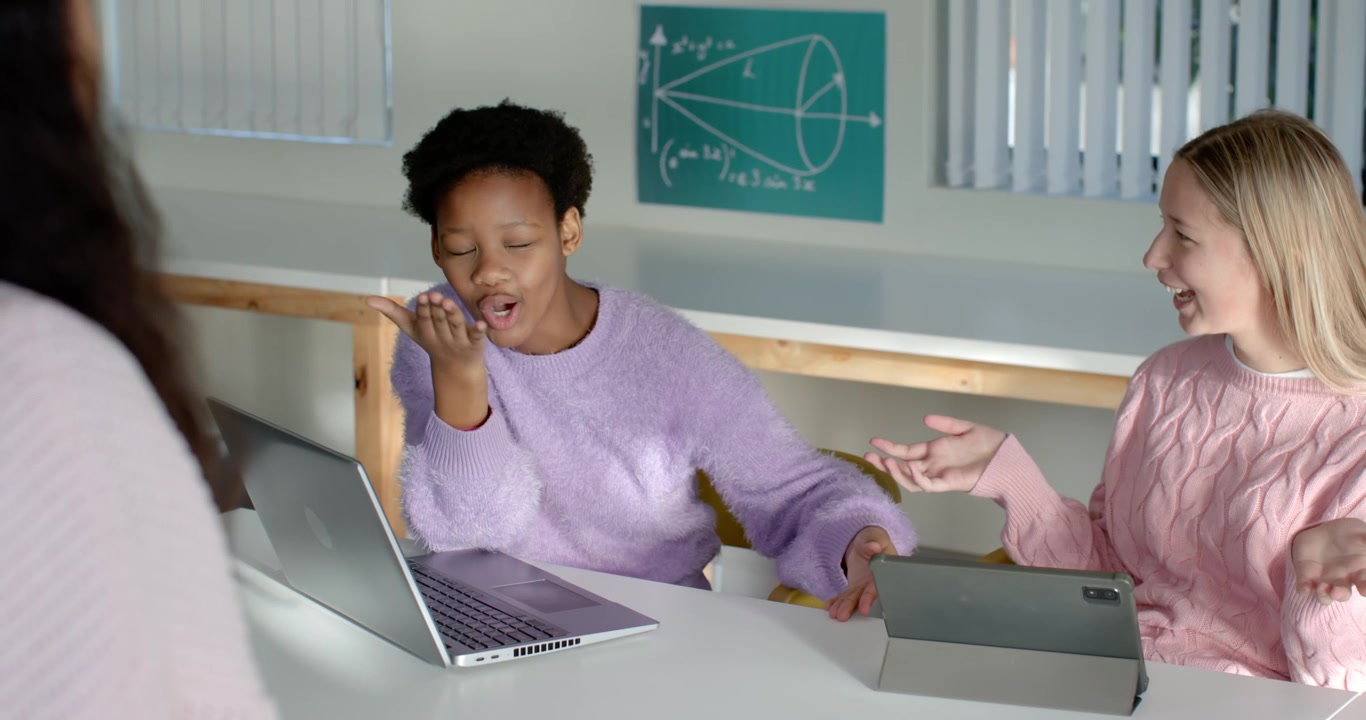 Teen females at table, lavender top gesturing to laptop, pink top mirroring tablet, discussing work