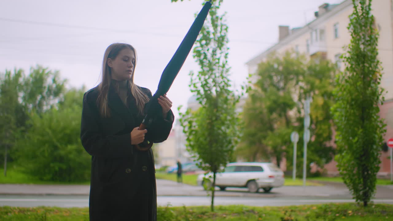 Woman in black trench coat holding closed umbrella looks up at cloudy sky with raised hand, standing outdoors on city street surrounded by greenery, cars, , anticipating possible rainfall