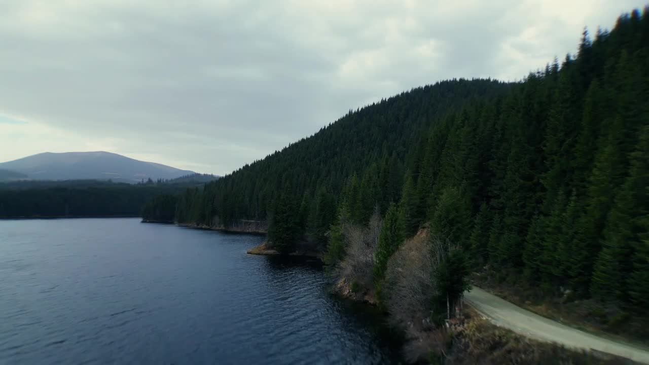 Drone flies along the shore of Oașa Lake near Oașa Dam in Romania, with dark blue water on the left, green forested bank on the right, cloudy day