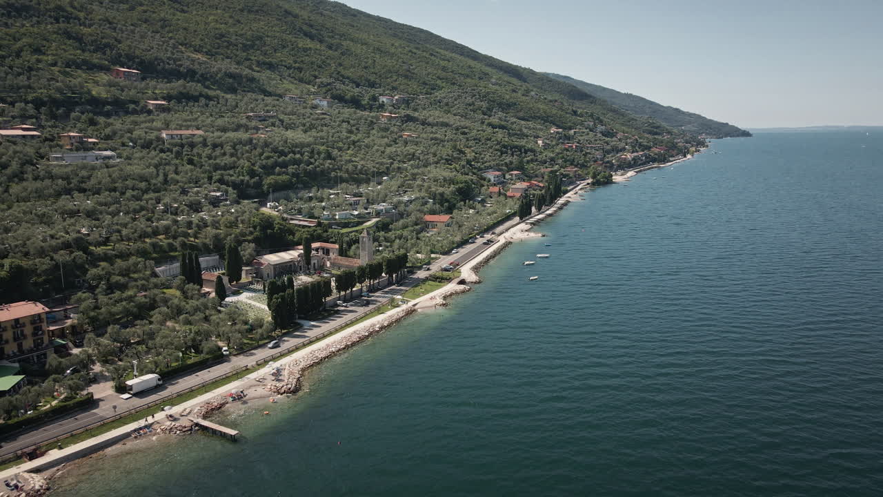 Aproximação cinematográfica de drone 4K UHD voando sobre a exuberante costa de Malcesine, Lago de Garda. Apresenta vilas com telhados vermelhos e água azul — ideal para viagens de luxo e anúncios imobiliários