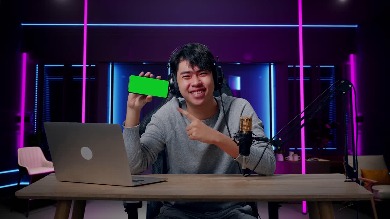 A Smiling Asian Man With Headphone And Computer Showing Green Screen Mobile Phone In Room Lit By Neon Lights In Retro