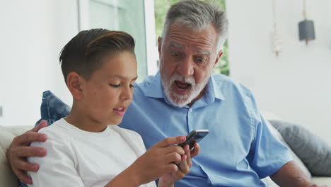 Nieto-Hispano-Mostrando-Al-Abuelo-Cómo-Usar-El-Teléfono-Móvil-En-Casa