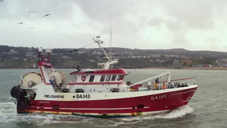 aerial sideview tracking follows fishing vessel in open ocean waters with flock of seagulls