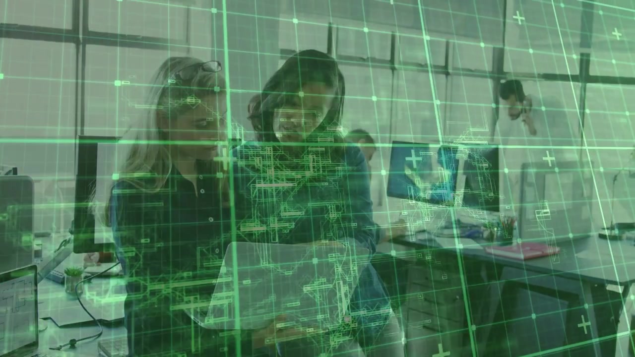 Two women holding laptop, one pointing, reviewing tech UI while green grid drifting across screens