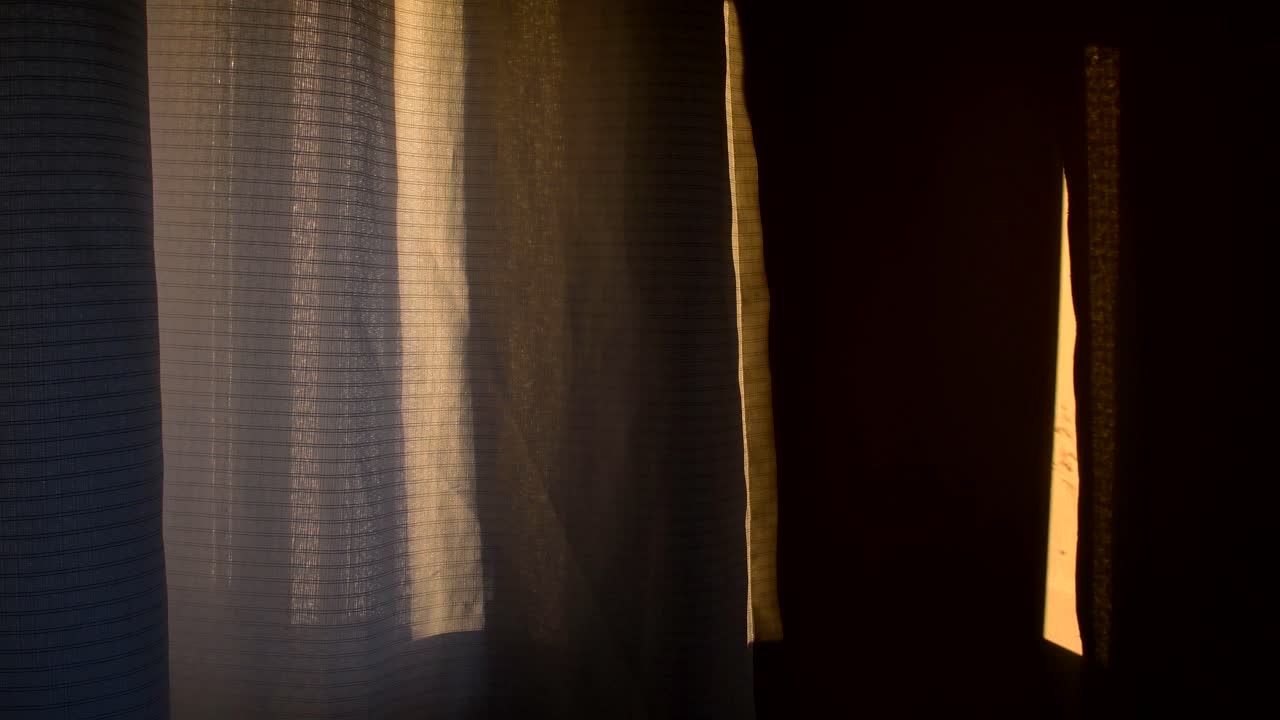 A curtain is moved slowly by the breeze, with warm dusk light falling on a nearby wall fading slowly to black