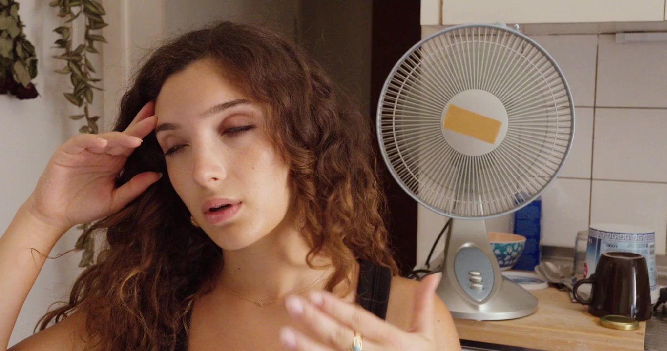 Žena si utírá čelo, protože se cítí přehřátá v interiéru, zatímco v pozadí běží elektrický ventilátor během horkého letního počasí.