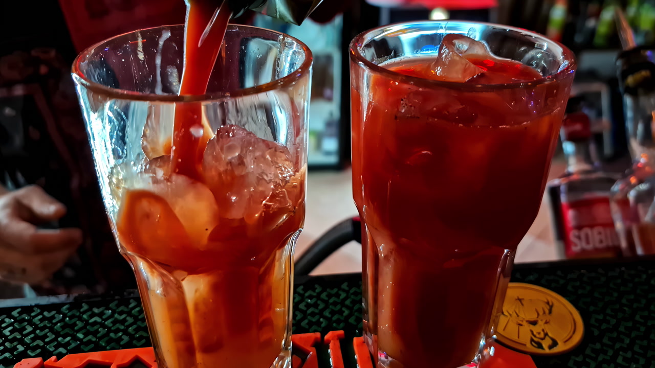 Bartender pours tomato juice into a tall glass beside an ice-filled drink on a bar mat. Close-up cocktail prep with splashes, refreshing brunch vibe, and nightlife setting.