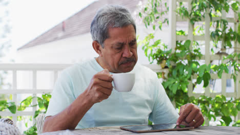 Senior-mixed-race-man-having-coffee-using-tablet-in-garden