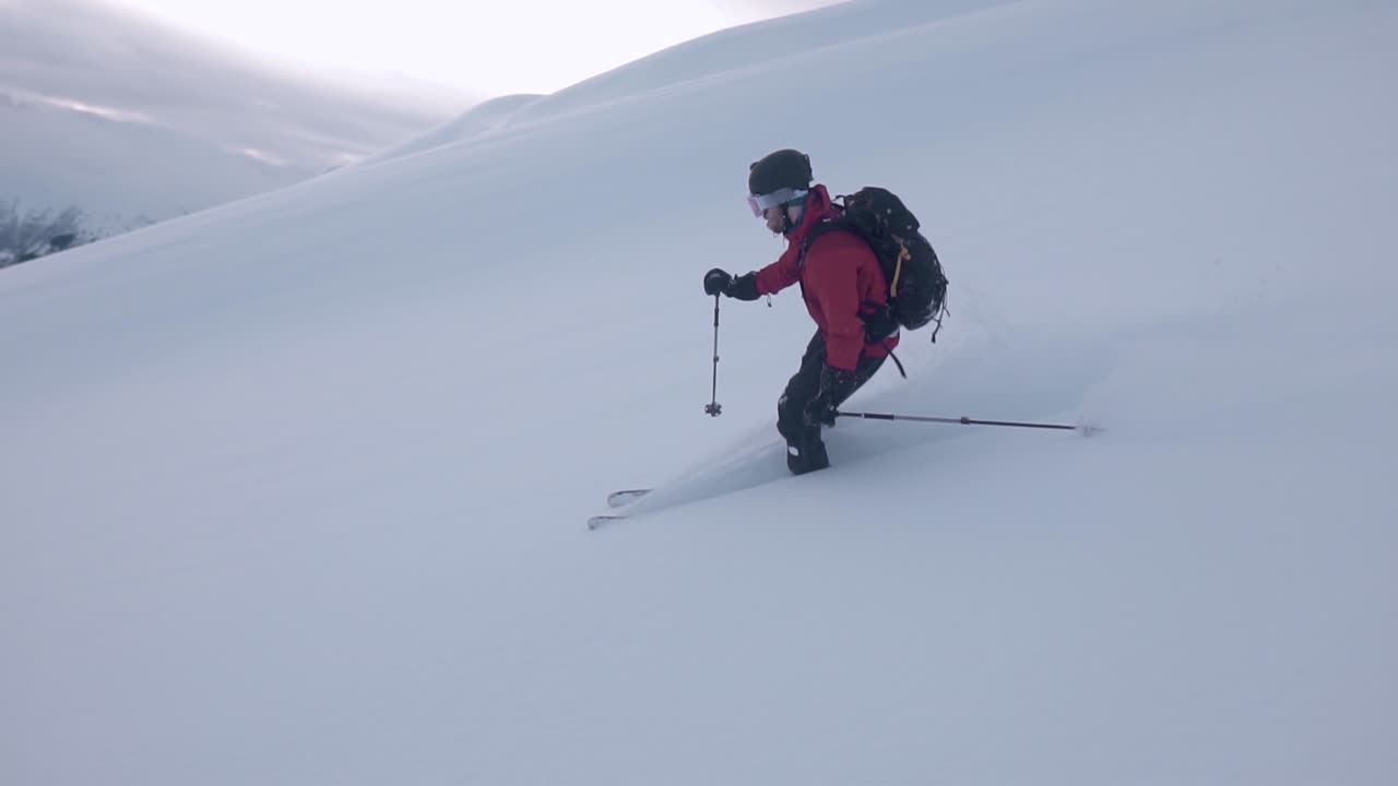 toma en cámara lenta de un hombre con chaqueta roja, esquiando cuesta abajo, rodeado por una montaña blanca como la nieve y un fondo semisoleado cubierto de nubes