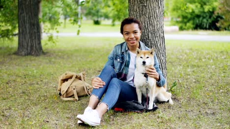 portrait of african american girl loving dog owner sitting in park on grass with her beautiful pet, smiling and looking at camera. loving animals and nature concept.