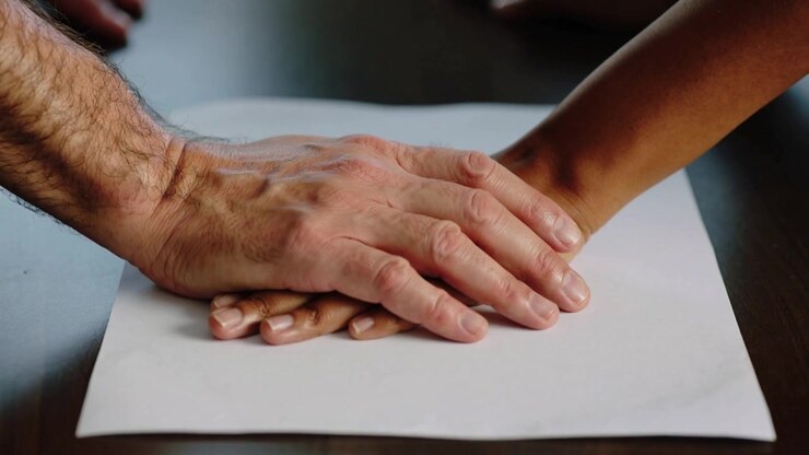 Approaching from left man's hand covering woman's hand on white paper at table for support lifting