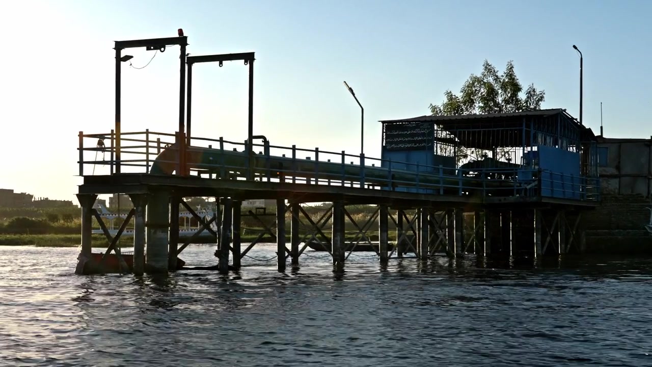 A view from a boat traveling on the Nile River, looking toward an industrial pier or water pumping structure elevated on stilts along the riverbank as sunset approaches.