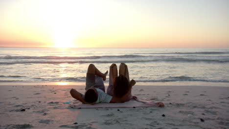 biracial couple relaxes on a beach at sunset, lying on a pink towel