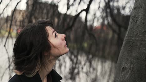 young woman in a black coat standing under a windy tree in a park in autumn in afternoon-1