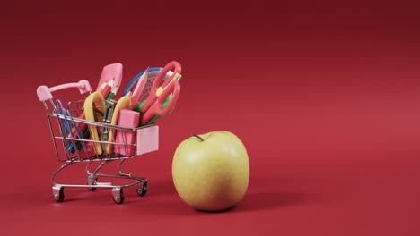 Close-up-of-shopping-trolley-with-school-items-and-apple-with-copy-space-on-red-background