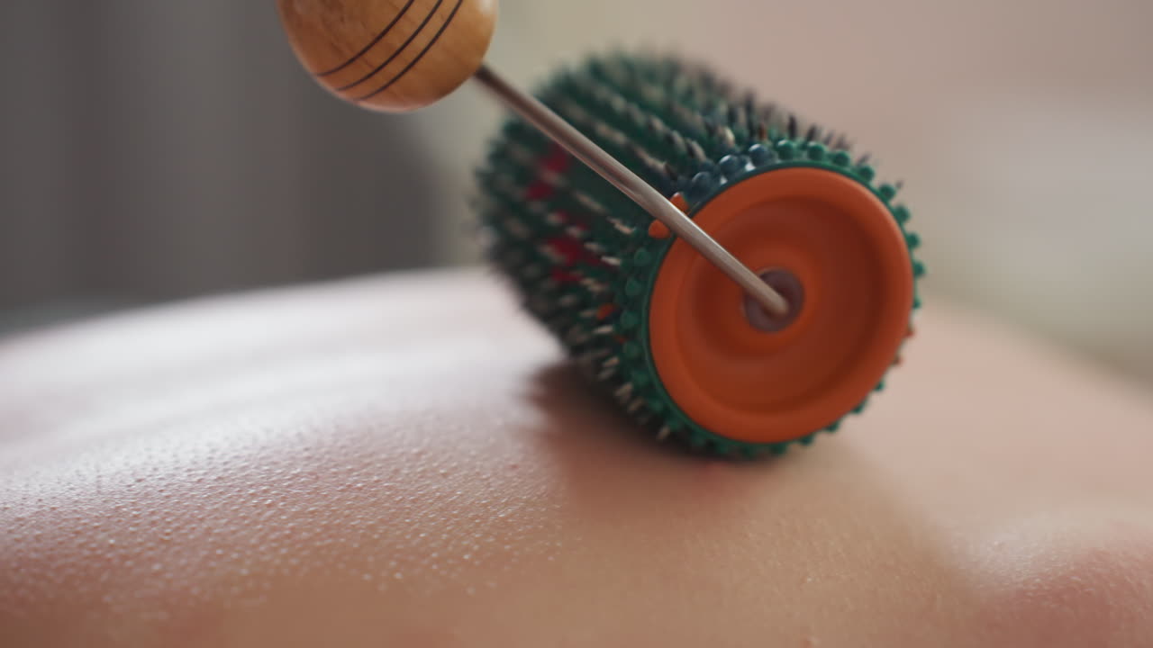 Close-up of colorful massage roller with green bristles, orange rim, and blue tips gliding across fair skin woman s back during relaxing treatment, showcasing detailed texture