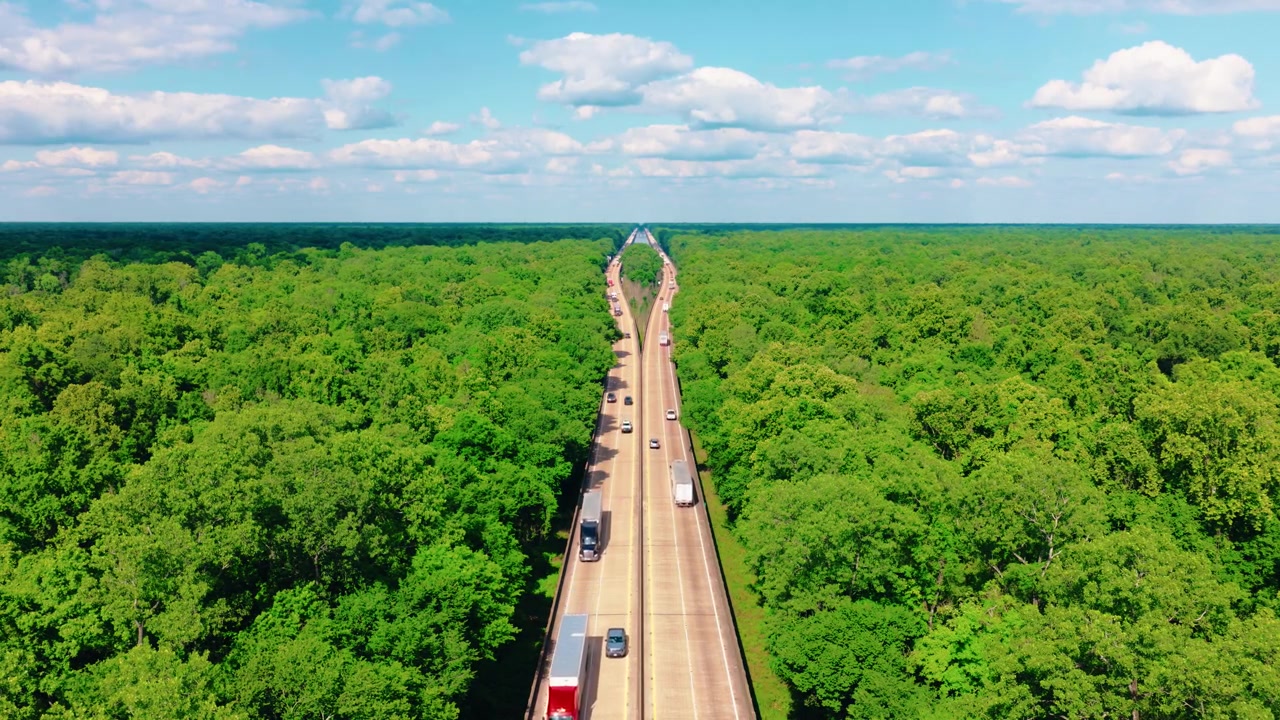 Pemandangan udara truk semi yang melaju di bentangan jalan layang I-10 melalui hutan Louisiana yang tak berujung di bawah langit musim panas yang biru — rekaman truk di jalan terbuka dekat Butte La Rose dengan lalu lintas hingga ke cakrawala.