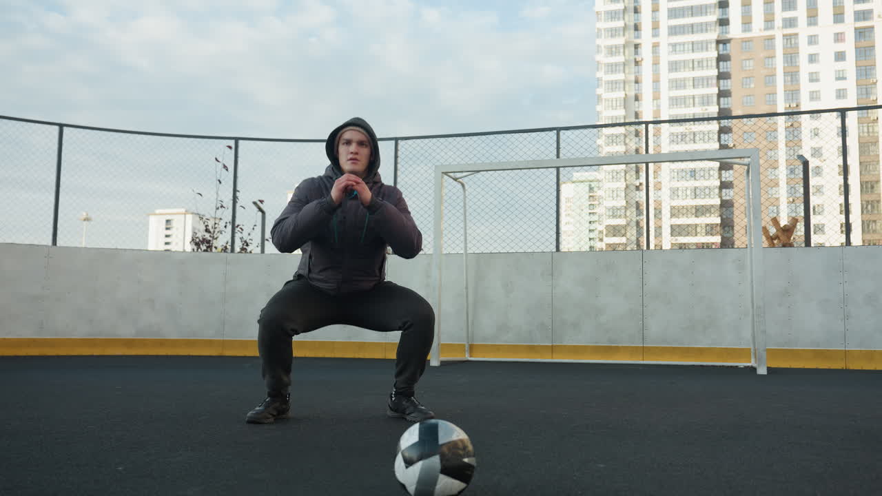Athlete performing deep squats with hands clasped in focus, preparing for exercise on outdoor sports ground with soccer ball in foreground, surrounded by urban high-rise buildings and goal post