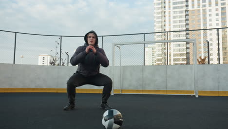 atleta realizando sentadillas profundas con las manos juntas en el foco, preparándose para el ejercicio en un campo deportivo al aire libre con una pelota de fútbol en primer plano, rodeado de edificios urbanos de gran altura y postes de gol