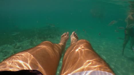 underwater personal perspective view of man legs floating in clear transparent sea water with people bathing in background, slow motion